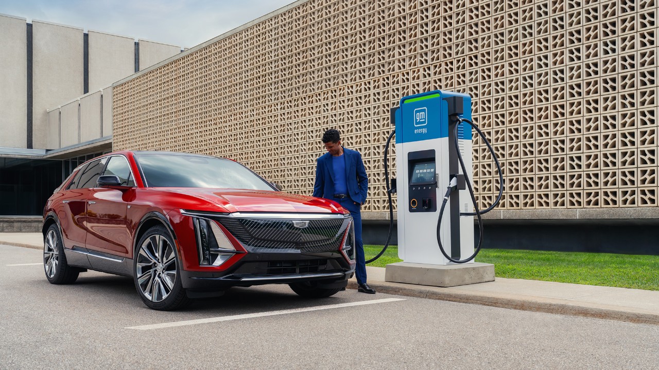 Public-Charging-Commitment A man in a blue suit standing by a red Cadillac LYRIQ electric SUV parked at a GM EVgo charging station.