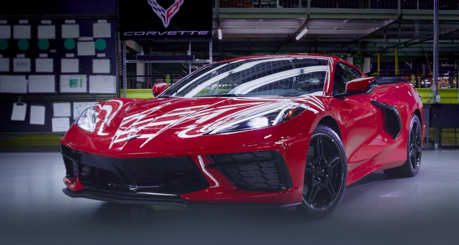 Close up of front /side of red Corvette sitting on shop floor with large Corvette Logo sign hanging on wall
