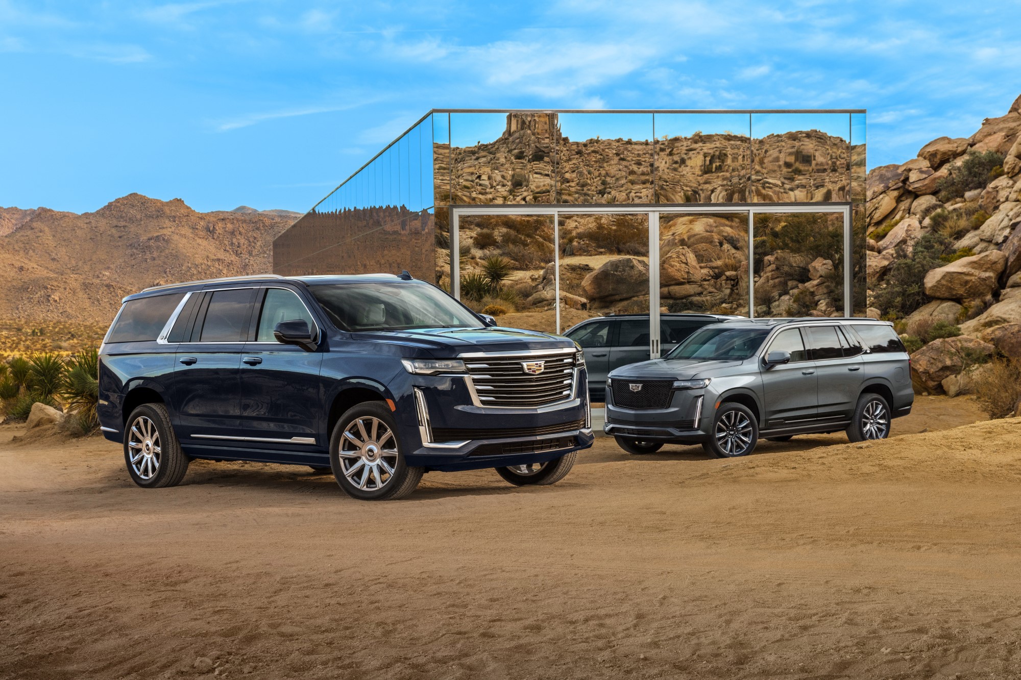 Front and side view of one black and one silver Cadillac Escalade ESV parked on dirt in front of a mirrored garage reflecting the rocky hills in the background.