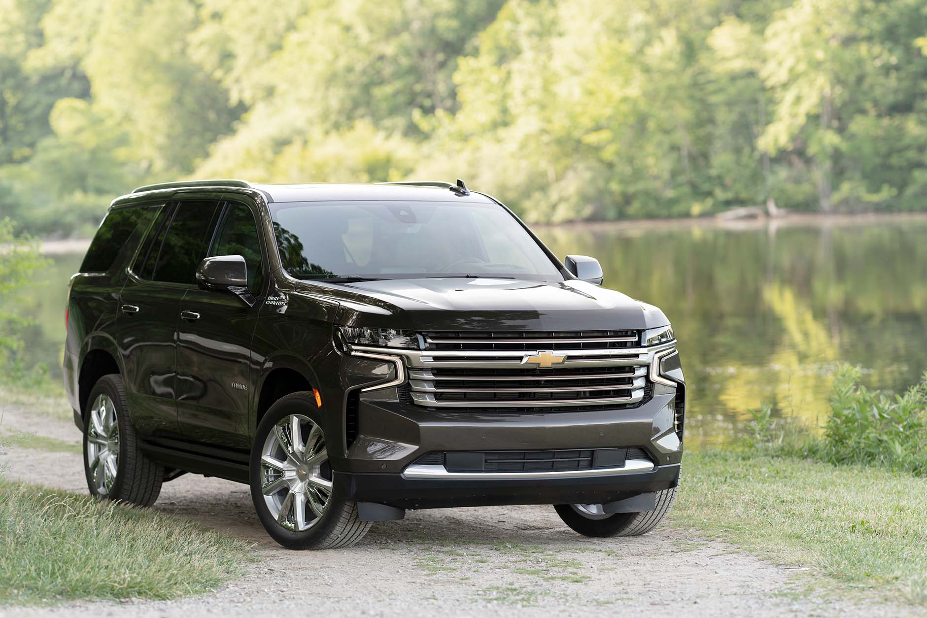 Front / side view of dark colored 2021 Chevrolet Tahoe High Country parked on sand driveway in front of lake and sun covered trees