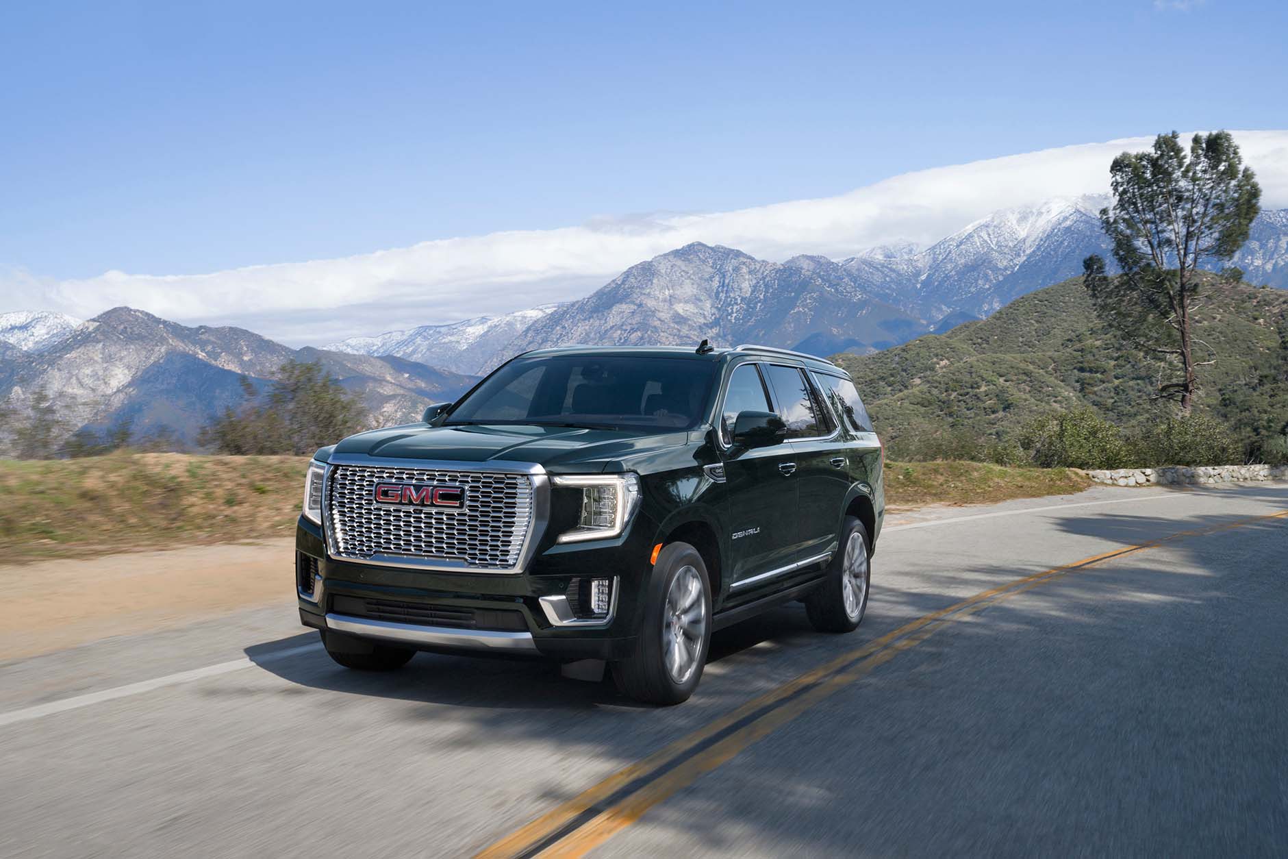 Front /side view of black 2022 GMC Yukon Denali driving down hilly road with snow-capped mountain peaks in the background