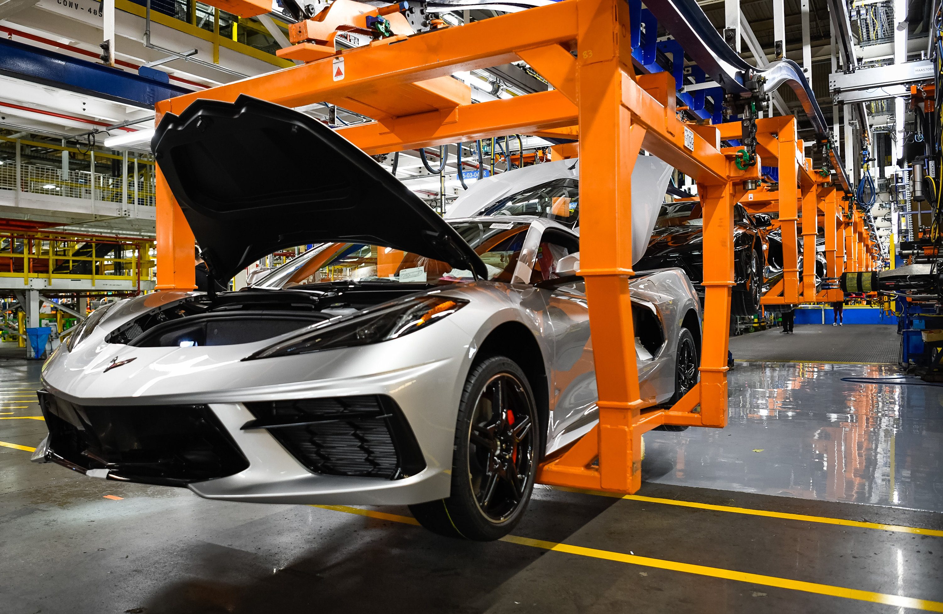 Orange lift moves silver corvette with hood and trunk open down the assembly line