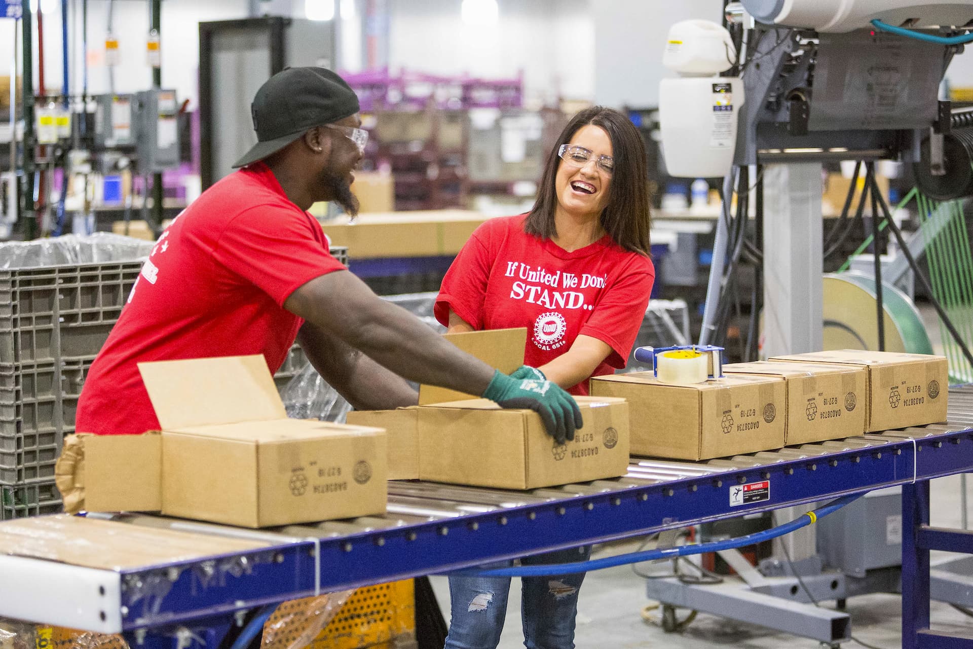 Two workers package auto parts in cardboard boxes for shipping.