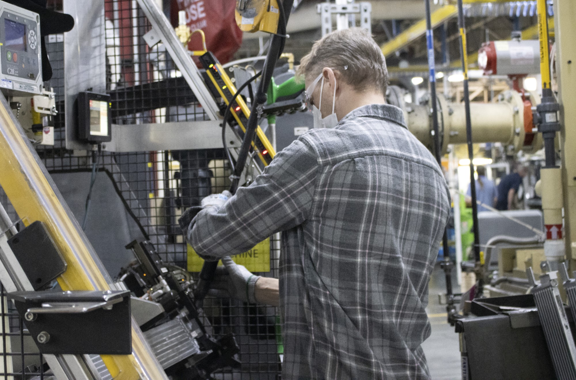 worker in PPE uses tool from over head to work on component on work station in front of him