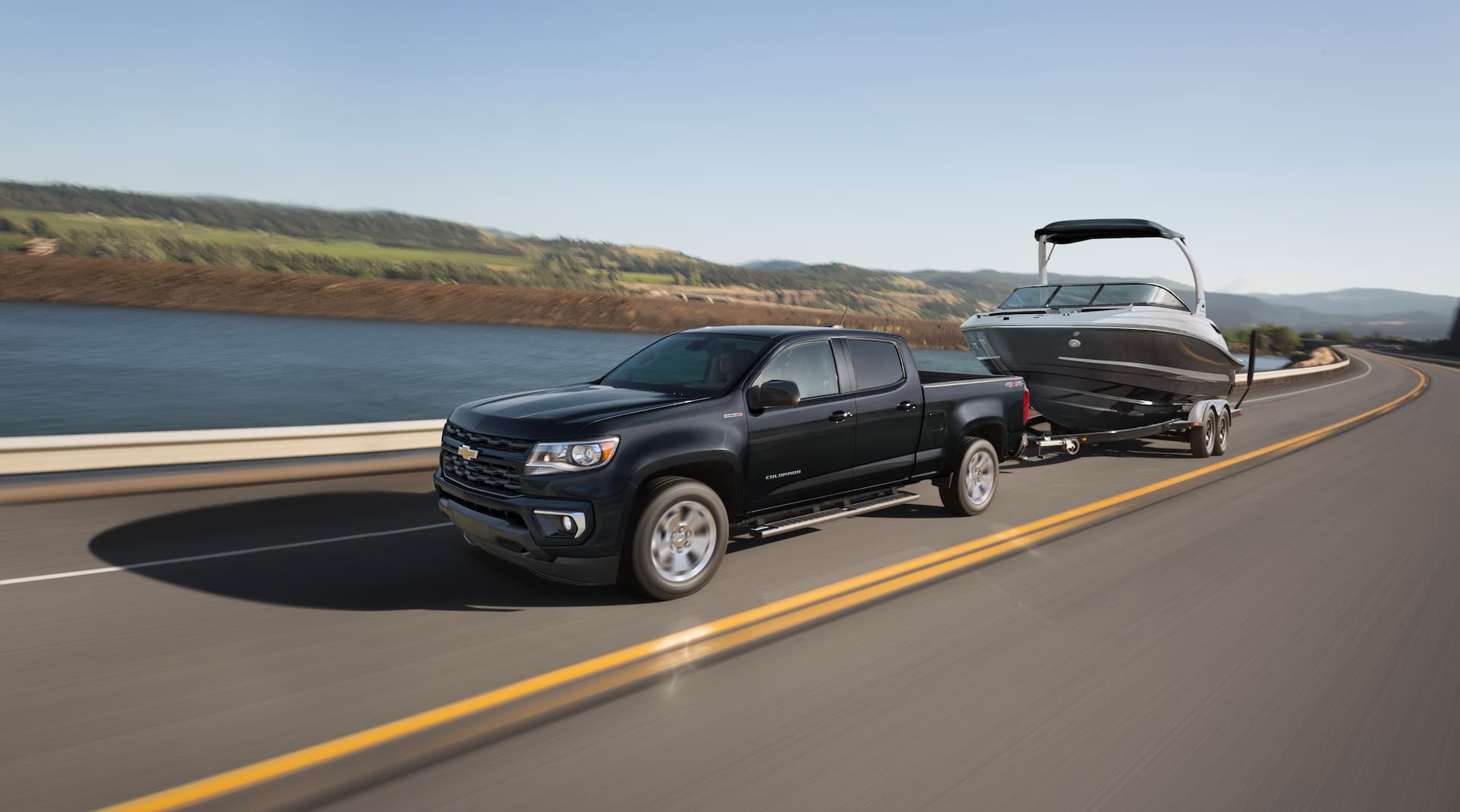 Black 2021 Chevrolet Colorado with dual cab tows a large boat down the road with large body of water In background