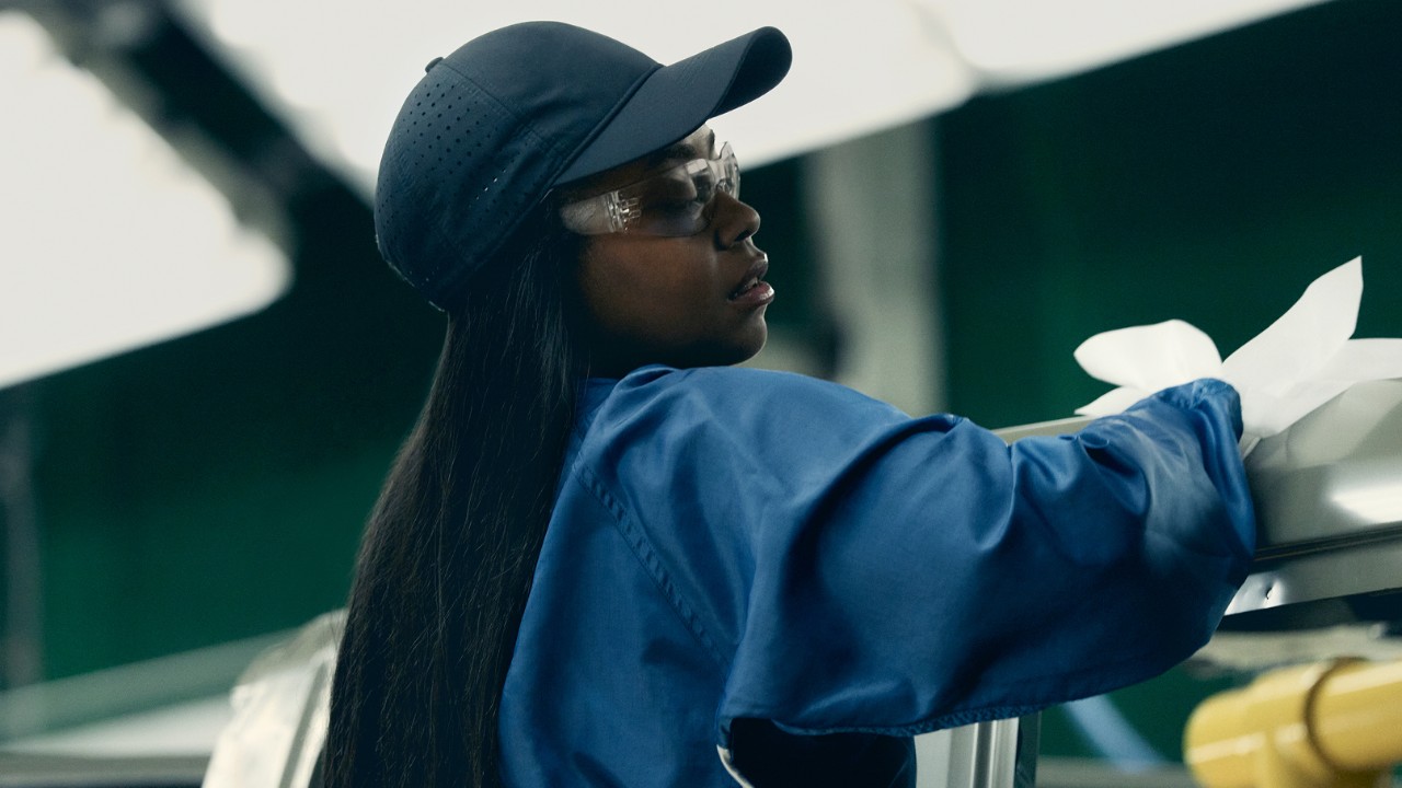 Drive Automotive worker wearing protective gear and inspecting a vehicle component in a factory environment.