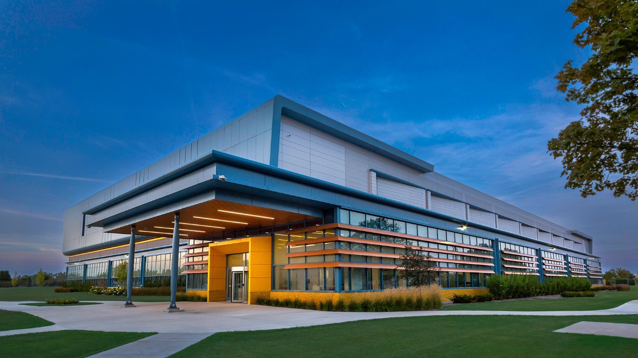 Modern facility with sleek blue, silver, and yellow exterior, surrounded by landscaped greenery at dusk.