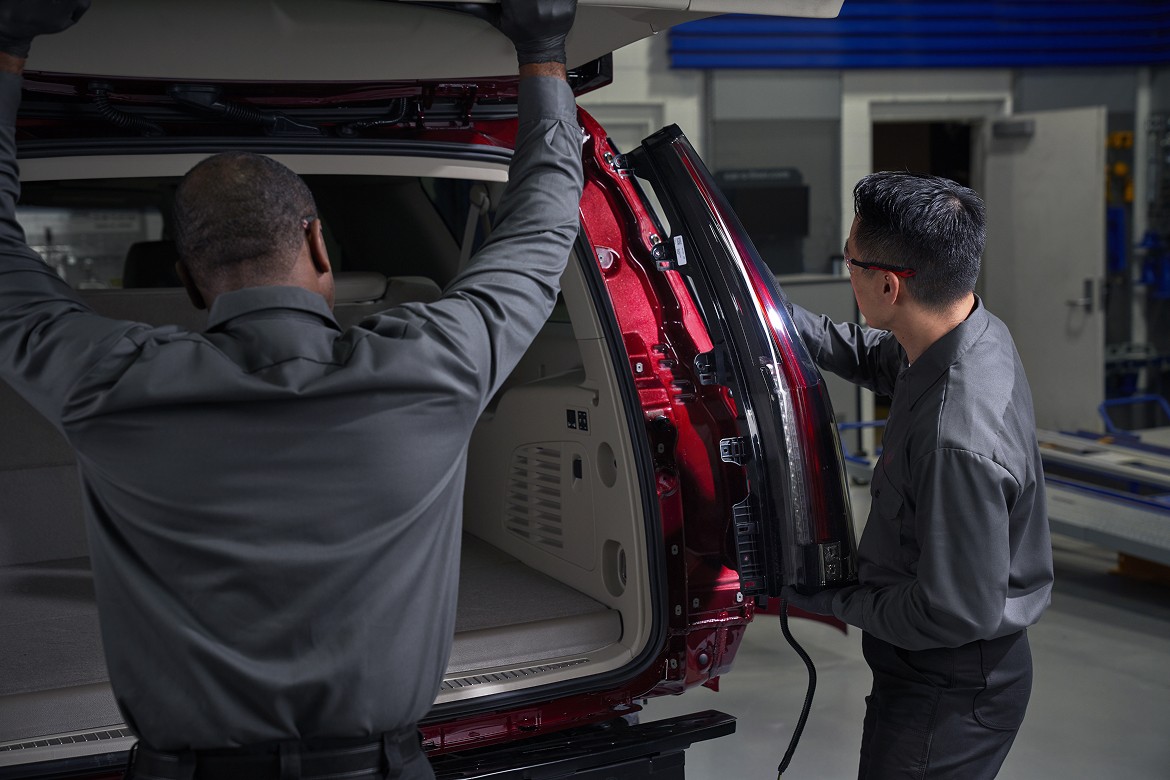 Two automotive technicians inspect the assembly of a red SUV with its rear liftgate open, inside a manufacturing facility.